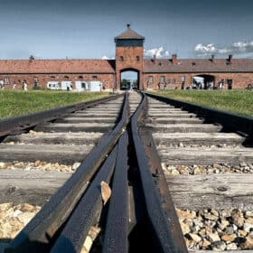 Dramatic perspective of the Auschwitz-Birkenau concentration camp railway, leading to the infamous entrance gate and watchtower under a bright blue sky. A sobering sight for visitors on the best Krakow to Auschwitz tours, invoking reflection on the tragedies of the Holocaust.