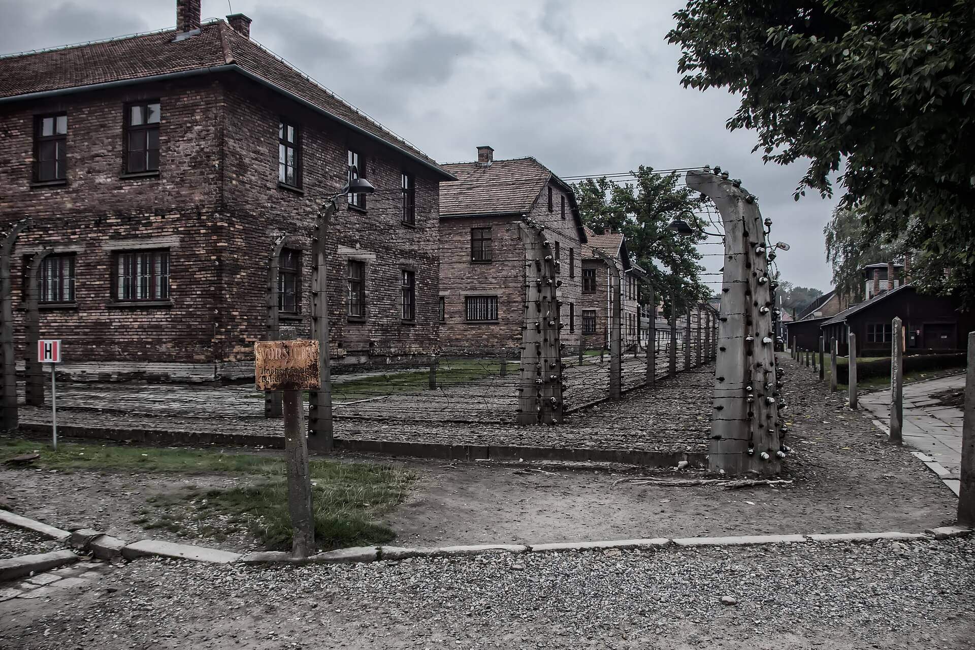 several two story buildings in a row at auschwitz with barbed ware fencing surrounding them and a old worn orange sign in German warning people to stay away-tours to auschwitz from warsaw