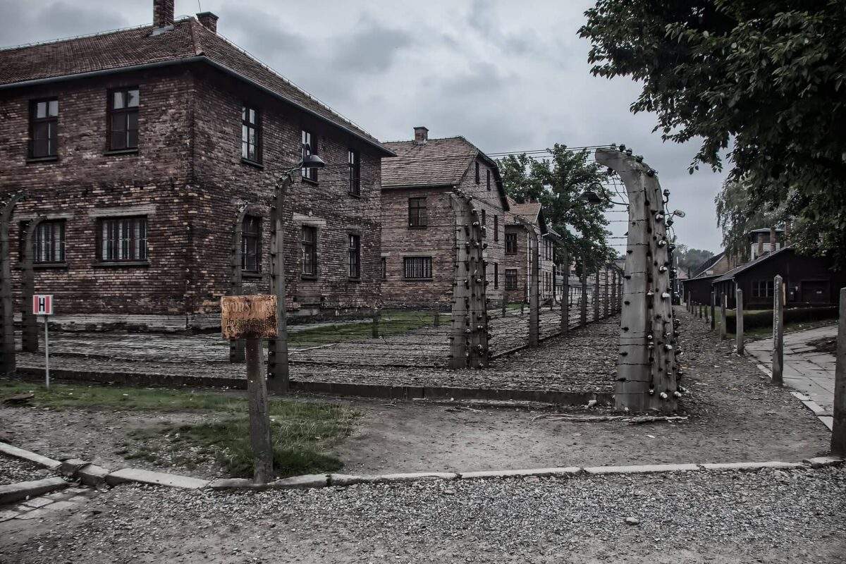 several two story buildings in a row at auschwitz with barbed ware fencing surrounding them and a old worn orange sign in German warning people to stay away