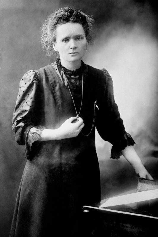 black and white image of marie curie with hair up, 3/4 sleeve dark dress, holding a necklace in her right hand, leaning against a table with her left hand neutral expression on face staring into camera