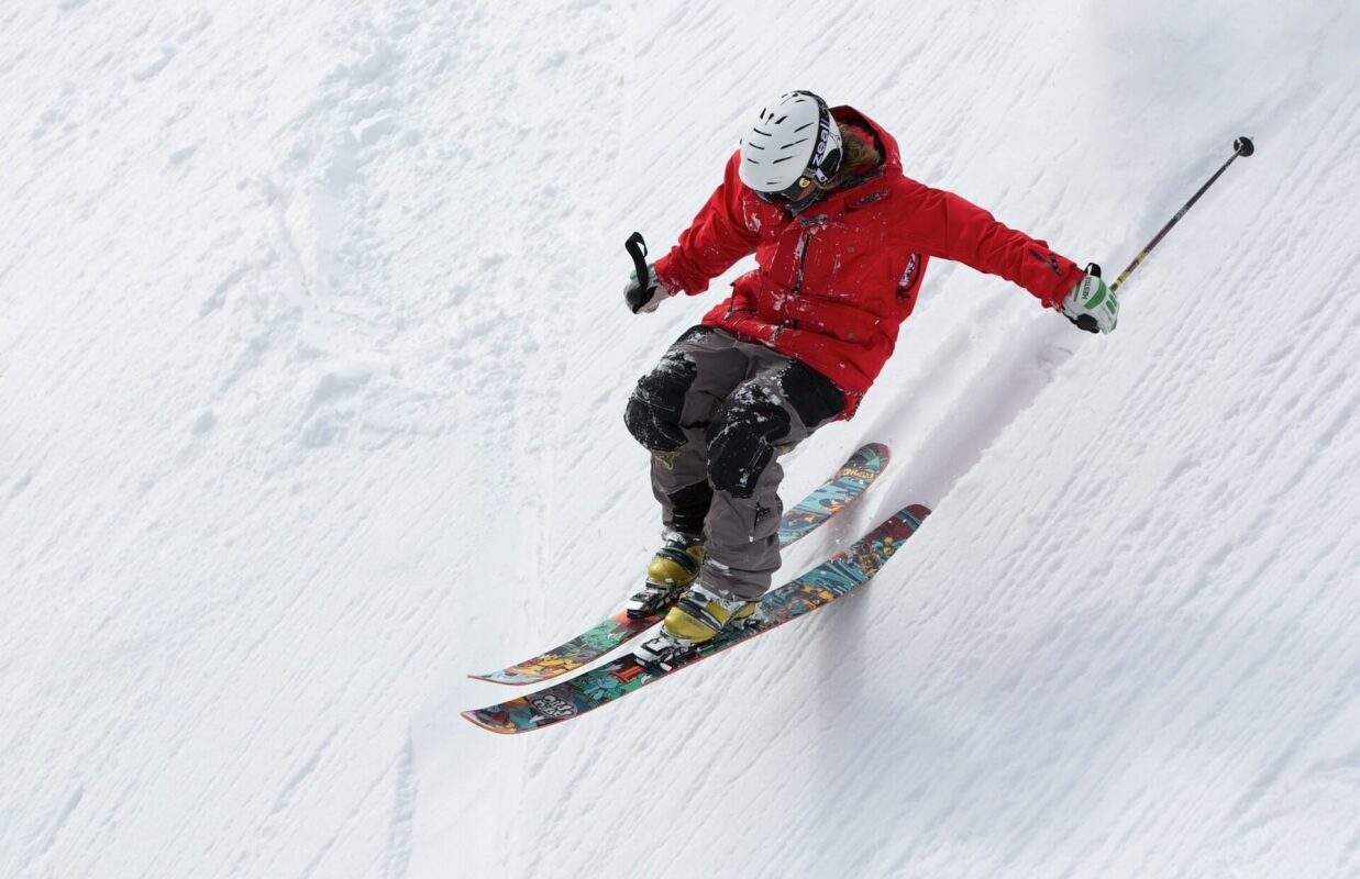 man in helmet, red ski jacket and black pants, on two skies with poles skiing down a deep slanted snowy hill
