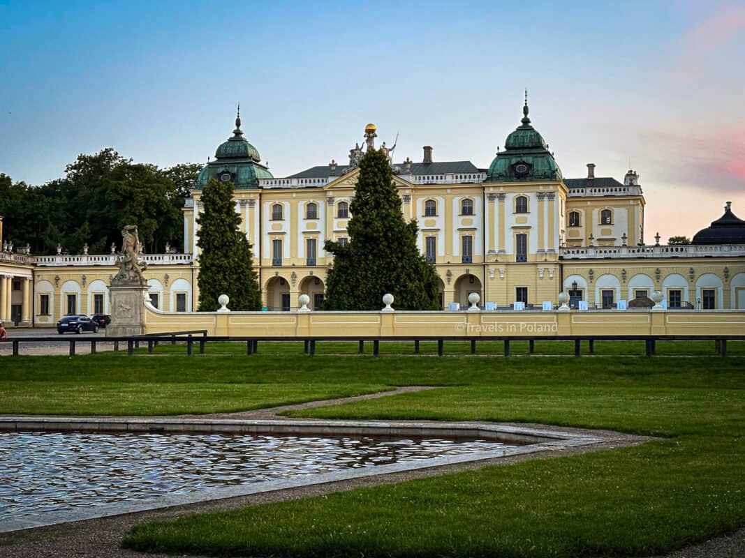 A grand view of the Branicki Palace in Białystok, Poland, featuring its classic baroque architecture with a pastel yellow facade and green domed roofs, set against a dusk sky. The image, with a watermark from 'Travels in Poland', captures the historical elegance that contributes to Białystok's reputation as one of the best cities in Poland.