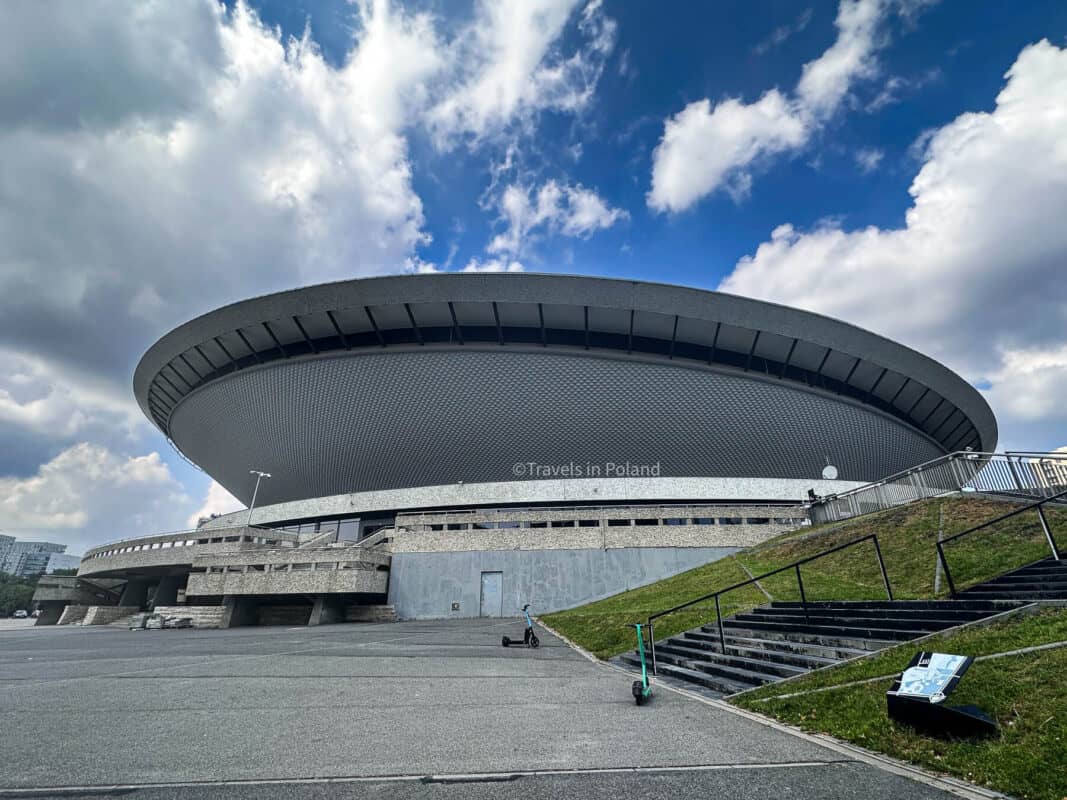 The imposing silhouette of the Spodek Arena in Katowice, with its distinctive saucer shape against a backdrop of dramatic clouds. This photograph, watermarked by 'Travels in Poland', underscores the modern architectural identity that places Katowice among the best cities in Poland for innovation and design.