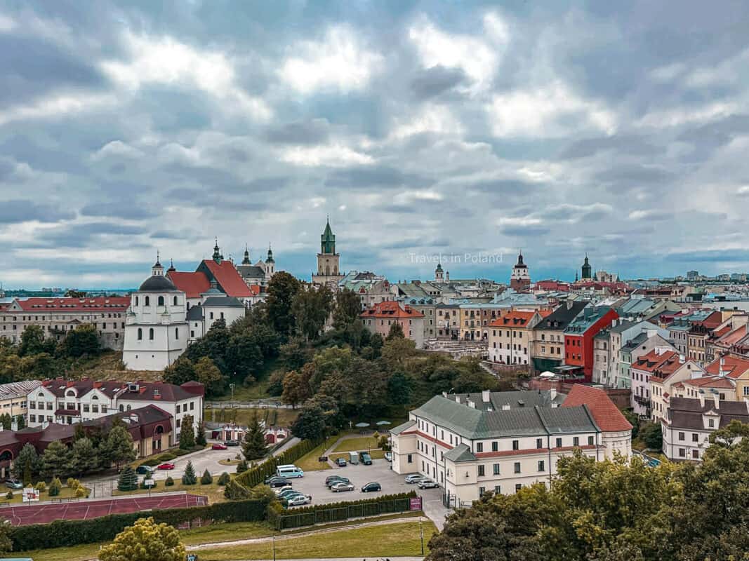 A panoramic view of Lublin's city center under a dramatic sky, showcasing a tapestry of historic buildings and church spires. This image, adorned with a 'Travels in Poland' watermark, highlights the cultural and architectural richness that ranks Lublin among the best cities in Poland