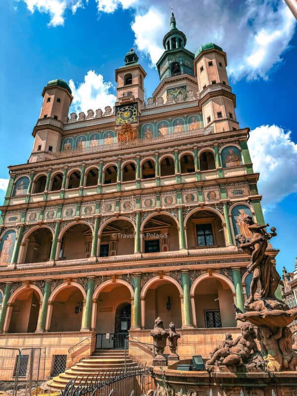 The Renaissance-style Poznań Town Hall, with its ornate clock tower and colorful façade, stands against a vivid blue sky with fluffy clouds. The Neptune Fountain, with its intricate sculpture, is prominently featured in front. This image, marked by 'Travels in Poland', captures the historic and cultural richness of Poznań, one of the best cities in Poland
