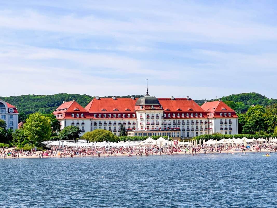 The Grand Hotel Sopot, an iconic white and red luxury establishment, stands by the sandy shores crowded with beachgoers under a clear blue sky. This picturesque scene captures the leisurely charm of Sopot, a coastal gem among the best cities in Poland, complete with the 'Travels in Poland' watermark.
