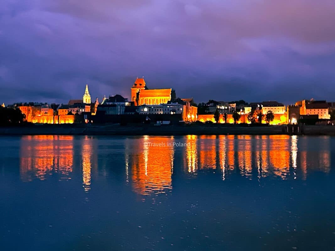 The medieval skyline of Toruń glows with warm lights against the twilight sky, mirrored beautifully in the Vistula River. This photograph, credited with 'Travels in Poland', showcases the historic allure of Toruń, renowned as one of the best cities in Poland.