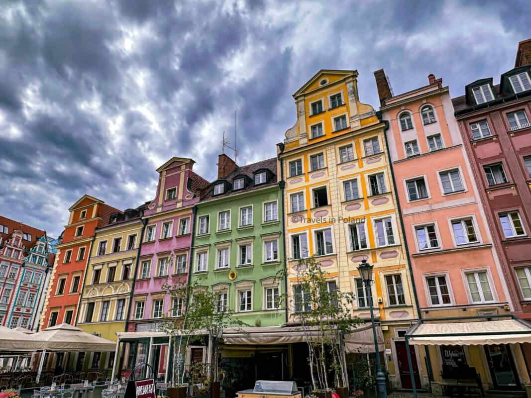 Colorful facades of historic townhouses in Warsaw's Old Town line a cobblestone square, set against a dramatic sky. The 'Travels in Poland' watermark acknowledges the photographer and the location as a highlight of one of the best cities in Poland.