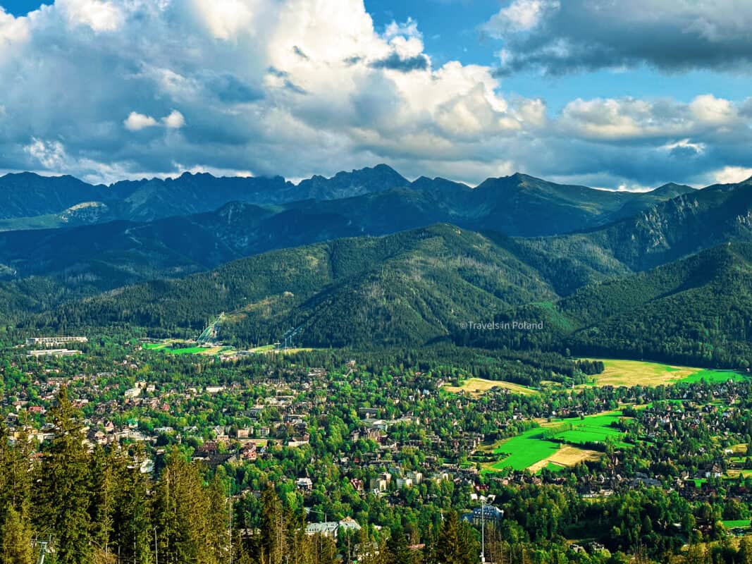 Overlooking the vibrant green landscape and dense forests of Zakopane, a picturesque mountain resort town nestled at the base of the Tatra Mountains. This aerial shot, credited with a 'Travels in Poland' watermark, highlights the natural beauty surrounding one of the best cities in Poland