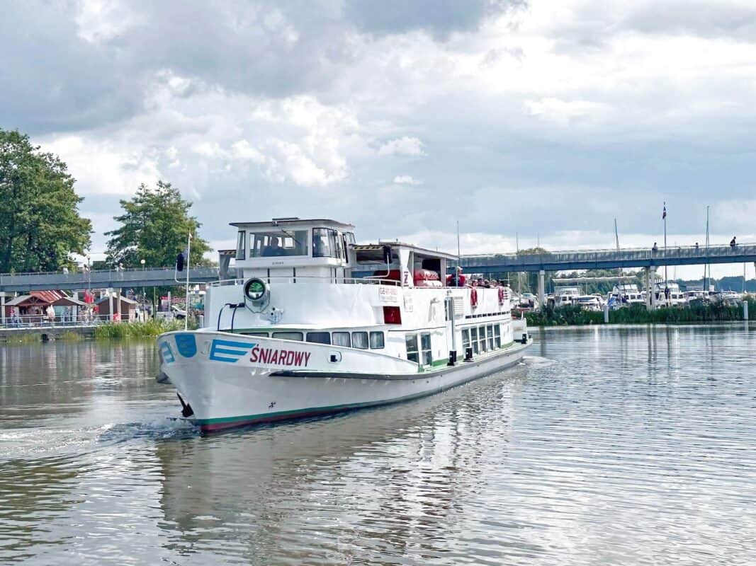 A white excursion boat named "Śniardwy" is sailing on a calm river under a cloudy sky. The background features a pedestrian bridge, trees, and people enjoying the riverside area. The scene captures a peaceful and leisurely moment.