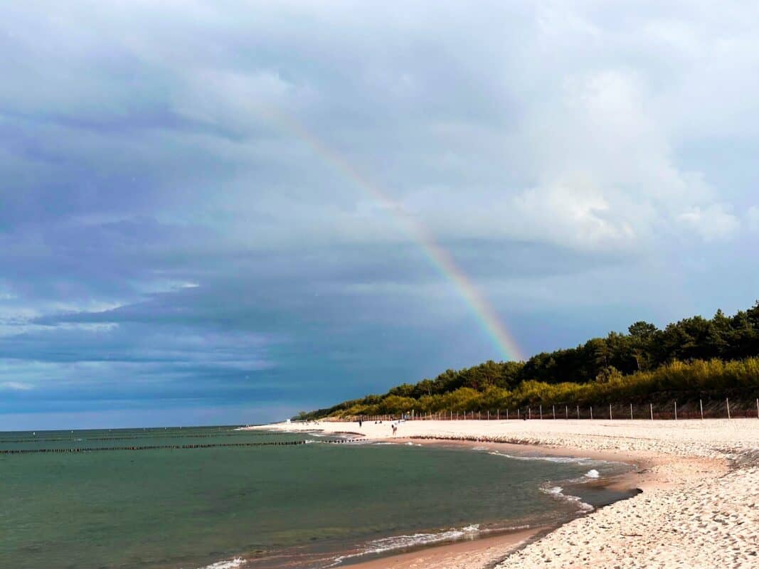 A scenic view of a sandy beach with gentle waves, bordered by lush greenery and a faint rainbow arcing across the cloudy sky. The peaceful shoreline stretches into the distance, creating a serene and picturesque coastal scene.