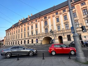This image shows a grand, historic building with a beige and yellow facade, lined with windows and a central arched entryway. Two cars, one silver and one red, are parked on the street in front of the building. The sky is clear and blue, and the scene is lit by the warm light of the setting or rising sun.